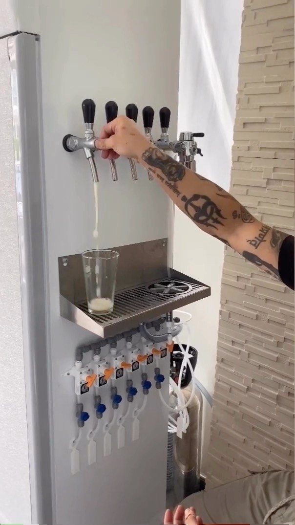 A tattooed arm pours a beverage from a silver beer tap into a glass.