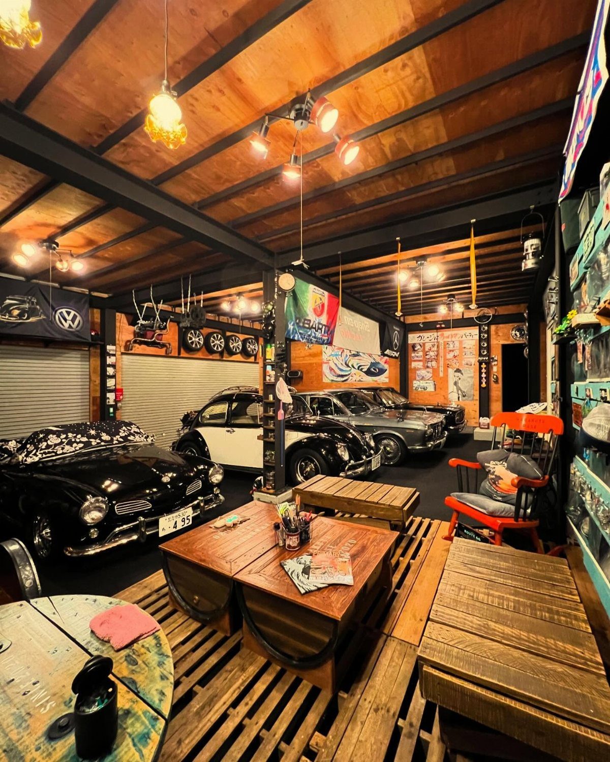 Classic cars parked in a luxury garage lounge with wooden pallet furniture and warm lighting.