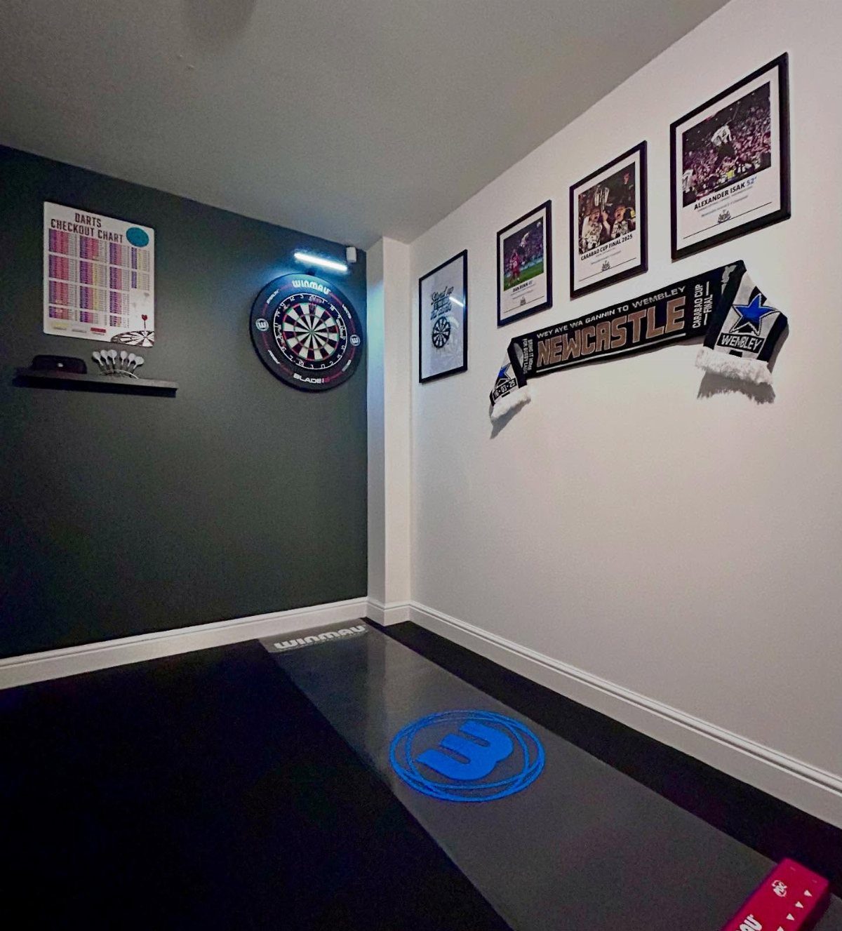 Dart board mounted on a dark wall next to a checkout chart and framed sports posters.
