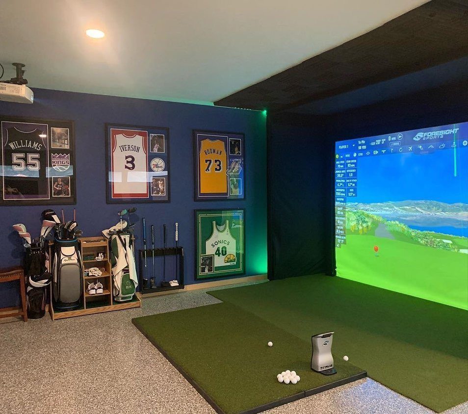 Golf simulator setup with a large projection screen, artificial turf, and sports memorabilia on blue walls.