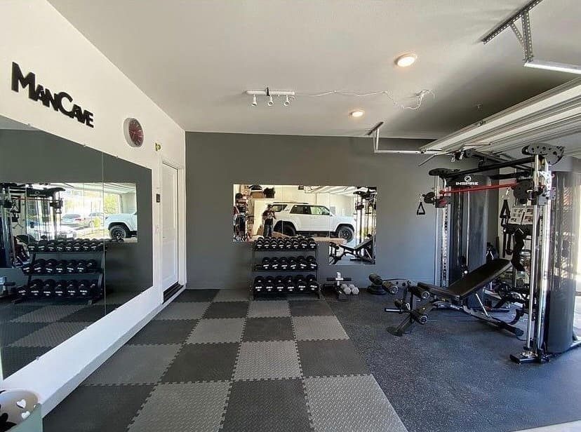 Home gym equipment including dumbbells, weight benches, and cable machines on a tiled garage floor.