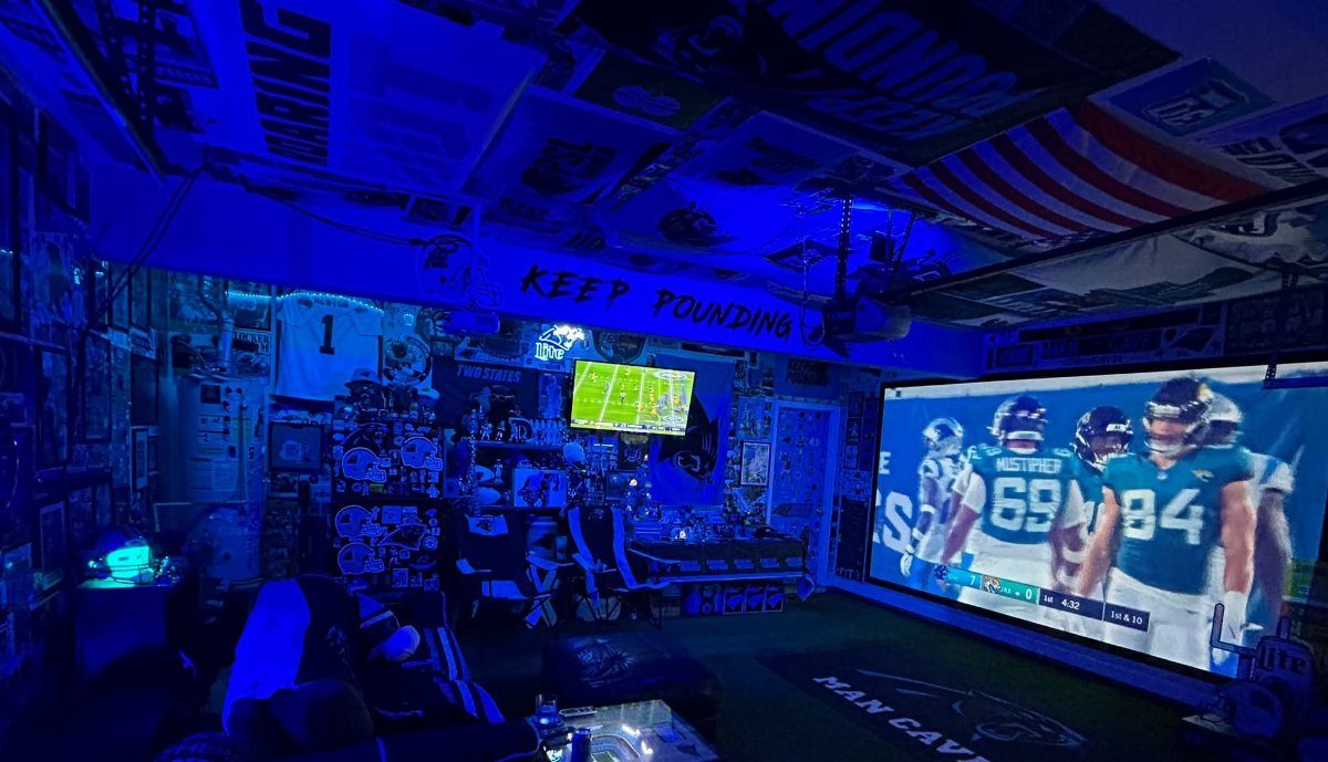 Large projection screen displaying a football game in a blue-lit garage man cave with sports memorabilia.