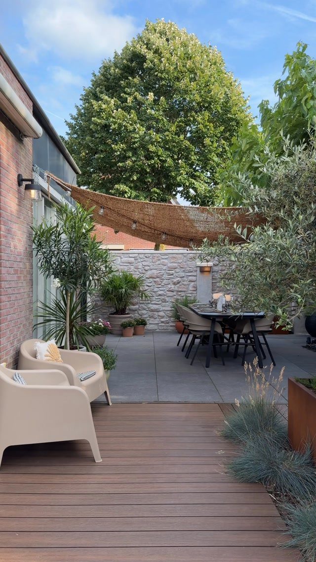 A modern patio featuring a wooden deck, stone paving, dining furniture, and a large woven shade sail.