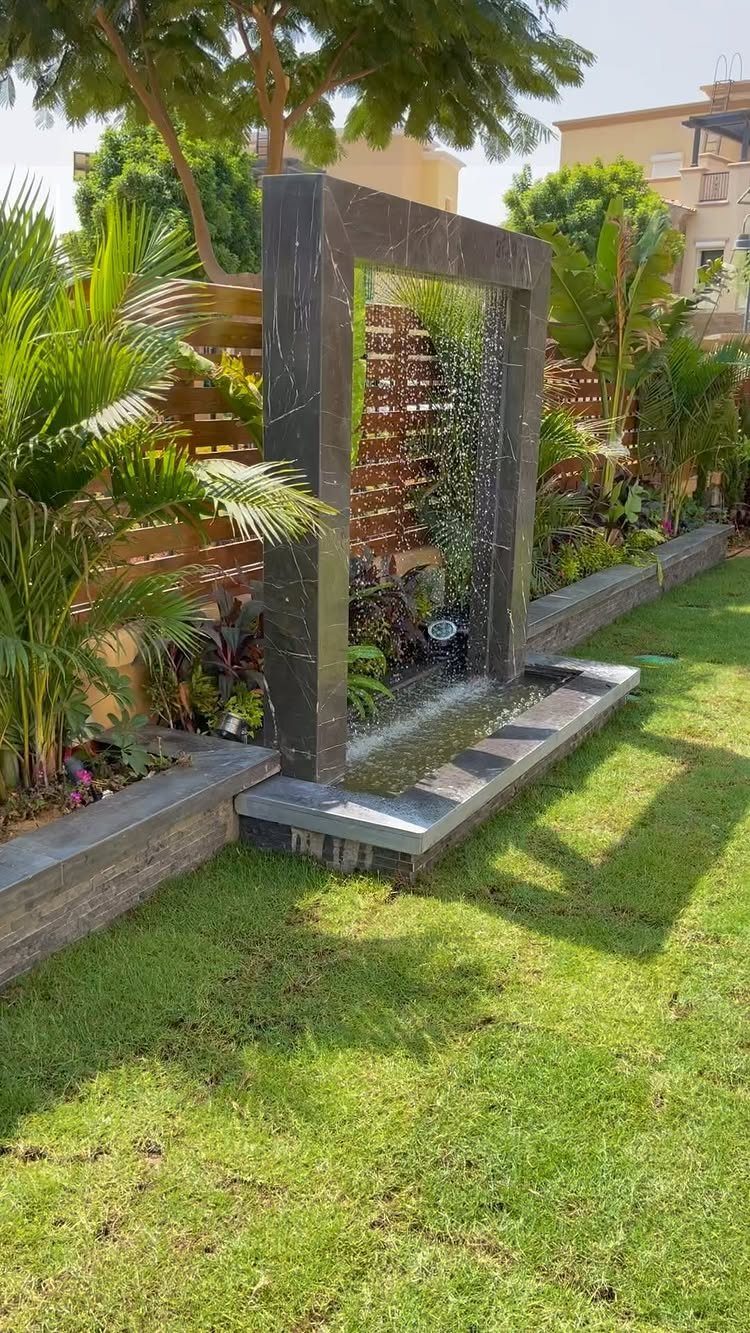 A modern dark stone water fountain cascading into a shallow basin in a green garden.