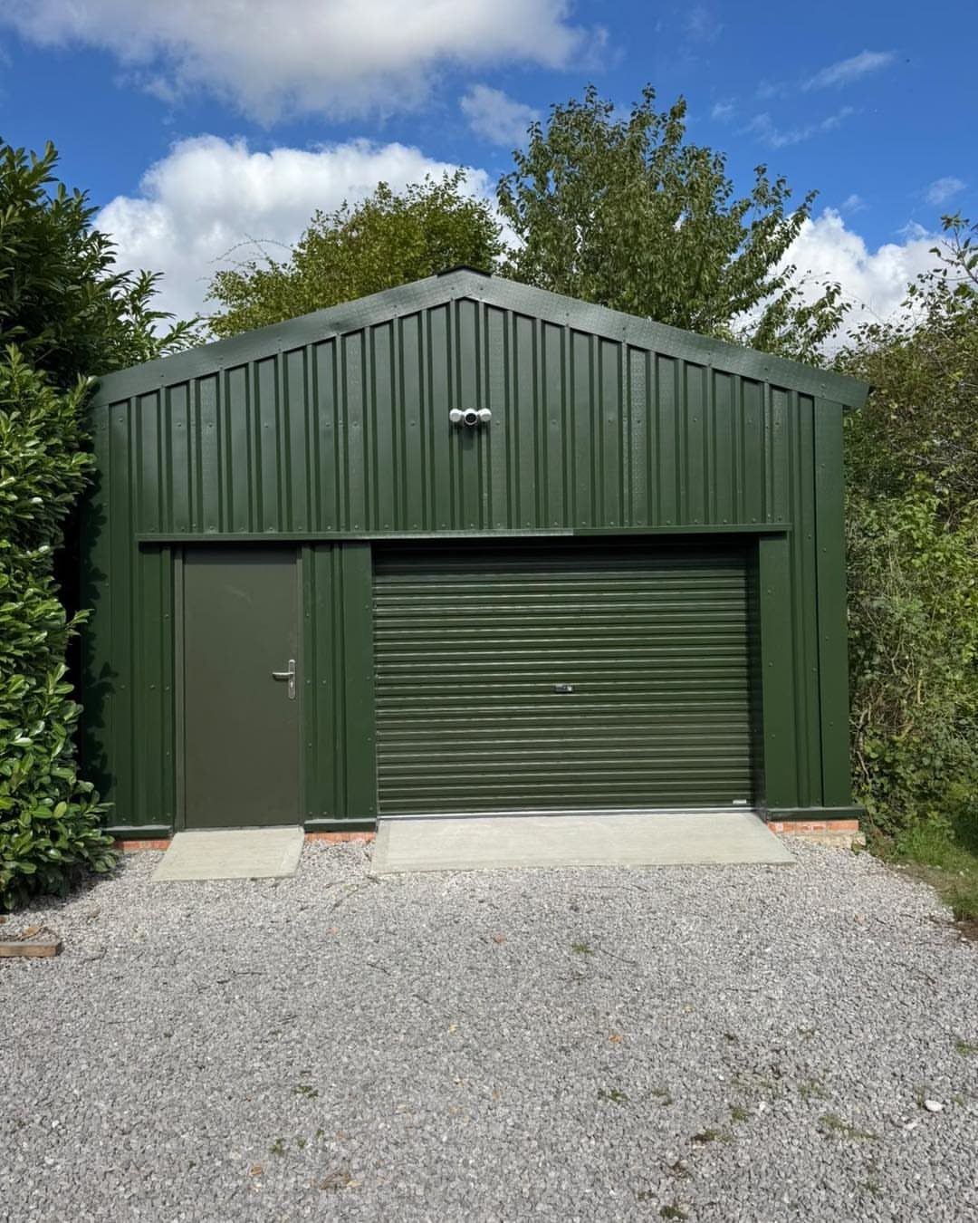 6 Painted Shed Colors for a Backyard That Actually Looks Planned 1 A large forest green metal garden building with a roller door and side door on gravel.
