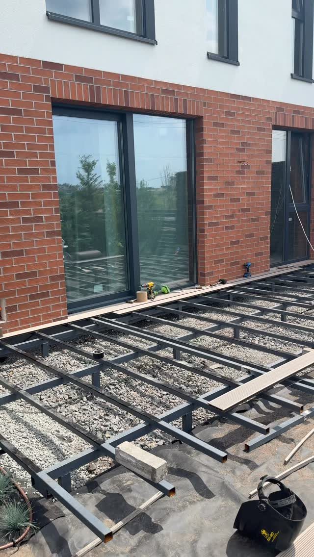 Raised Deck Frame: 8 Ideas From Wood Joists to Galvanized Steel 8 A grey metal raised deck frame being installed on gravel next to a brick house.